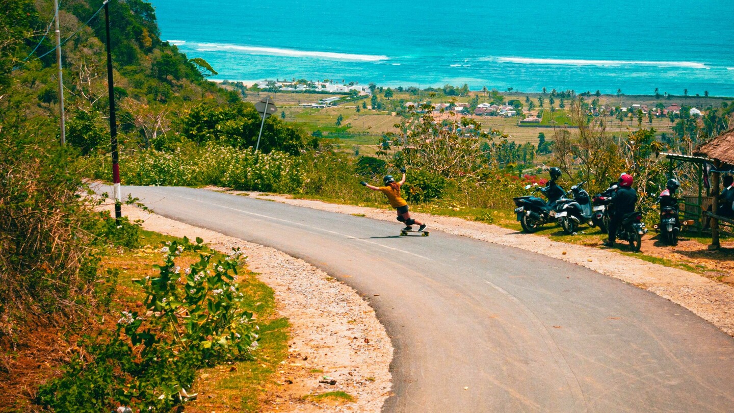 Eine Downhill-Skateboarderin fährt mit hoher Geschwindigkeit eine kurvige Strasse entlang, die zu einem herrlichen Küstenpanorama mit türkisblauem Meer führt. Die Strasse schlängelt sich durch eine grüne Landschaft, gesäumt von Vegetation und vereinzelten Motorrädern am Strassenrand. Im Hintergrund erstreckt sich der Ozean unter einem strahlend blauen Himmel mit weissen Wolken, was der Szene eine sommerliche und abenteuerliche Atmosphäre verleiht.