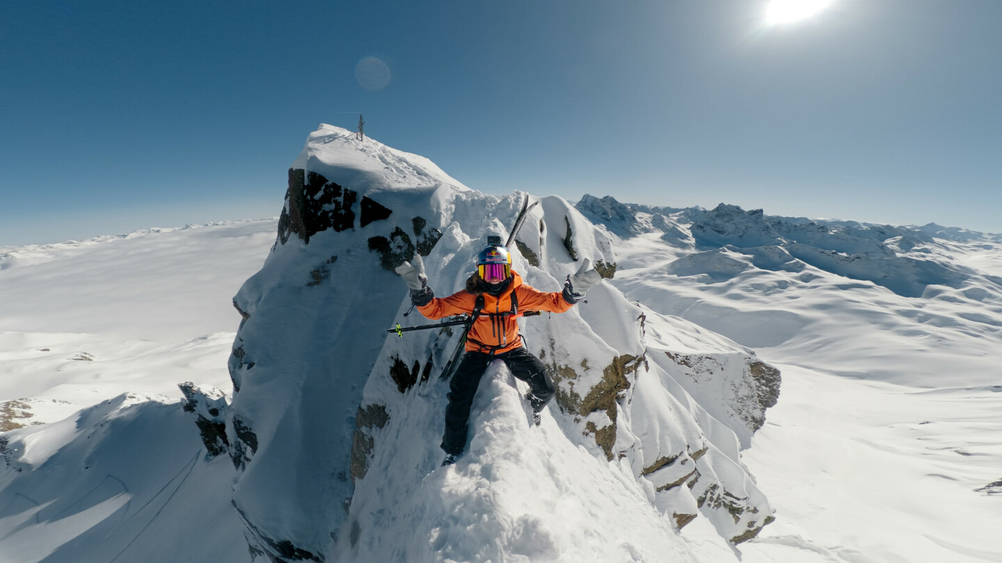 Skifahrer in orangefarbener Jacke balanciert auf einem schmalen, verschneiten Felsvorsprung, umgeben von einer spektakulären Winterlandschaft unter strahlendem Himmel.