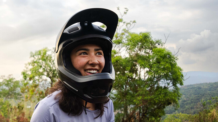 Samantha Soriano smiling while wearing a black full-face helmet outdoors. In the background, trees and a forested hilly area are visible, with a slightly cloudy sky.