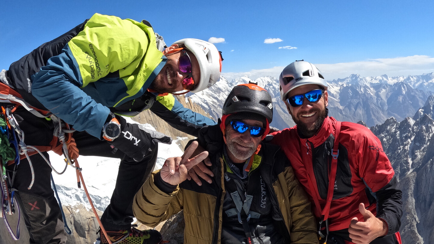 Trois alpinistes, dont Eduard Marín, sur un sommet enneigé avec équipement d'escalade et casques, souriant devant un paysage montagneux spectaculaire.