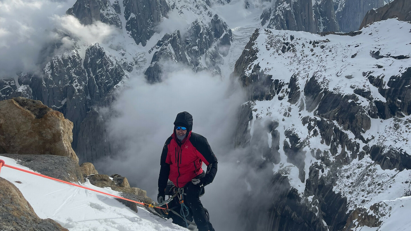 Climber in a red jacket ascending a snowy slope, surrounded by dramatic clouds and rugged mountain peaks.