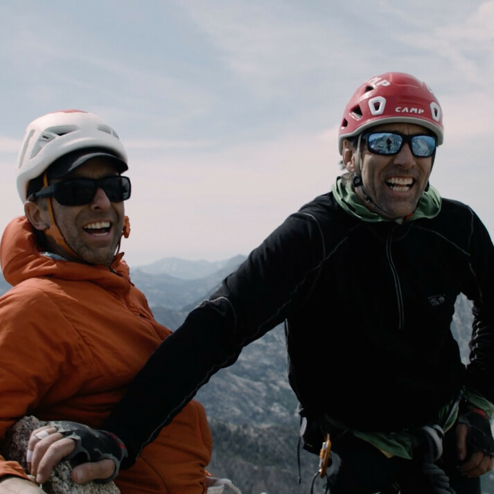 À gauche se tient Timmy O’Neill avec une veste orange et un casque, souriant avec des lunettes de soleil. À sa droite, le grimpeur aveugle Erik Weihenmayer porte une veste noire et un casque rouge. Les deux hommes se tiennent sur un sommet de montagne, heureux, profitant du vaste paysage montagneux en arrière-plan. L'image reflète l'aventure, l'escalade et la motivation.