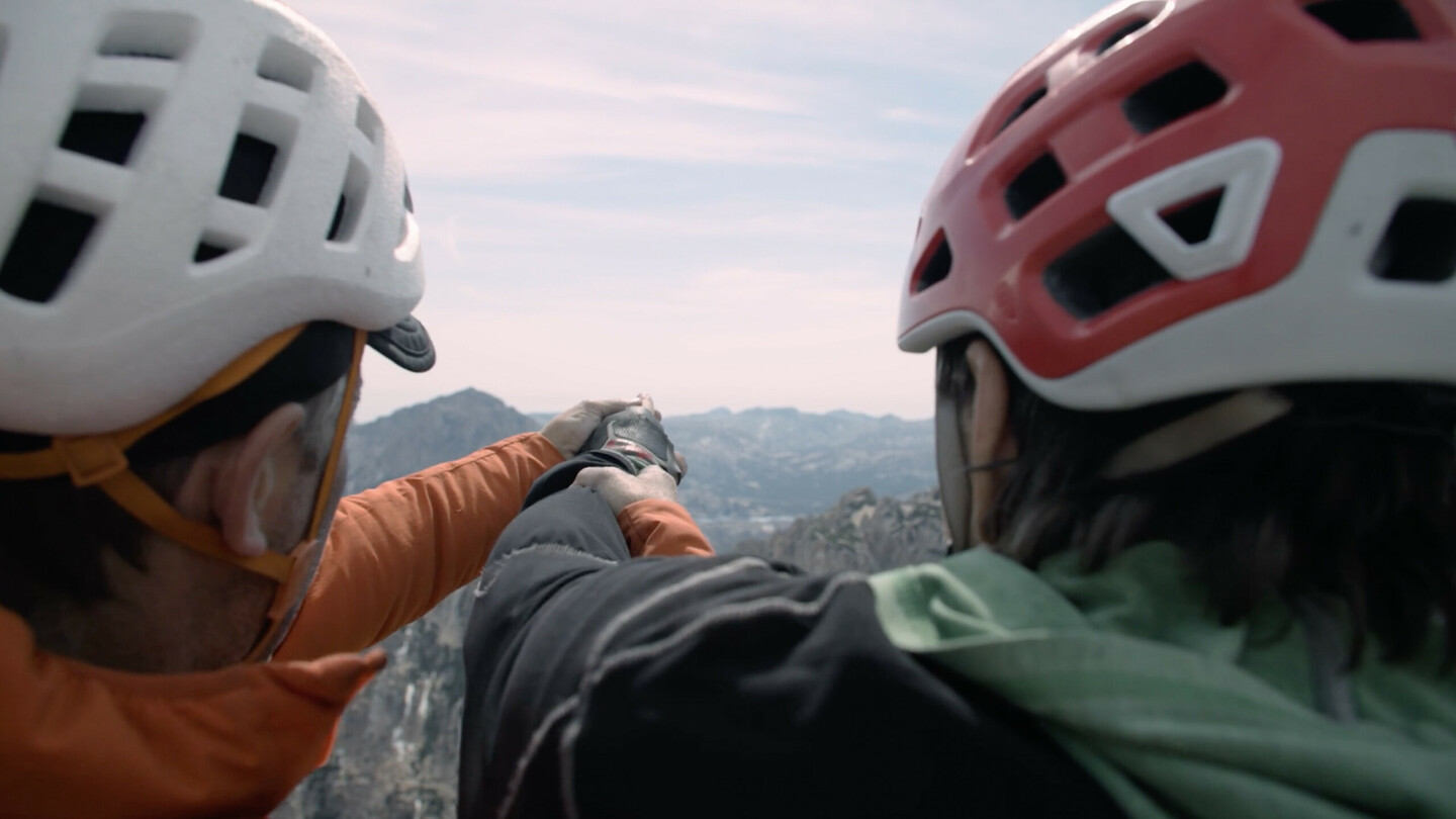 Gros plan sur Timmy O’Neill (à gauche) et Erik Weihenmayer (à droite) portant des casques, alors que Timmy guide la main d’Erik pour lui montrer le paysage montagneux. La scène met en avant l’esprit d’équipe et la collaboration solidaire entre les grimpeurs, avec les vastes sommets environnants en arrière-plan.