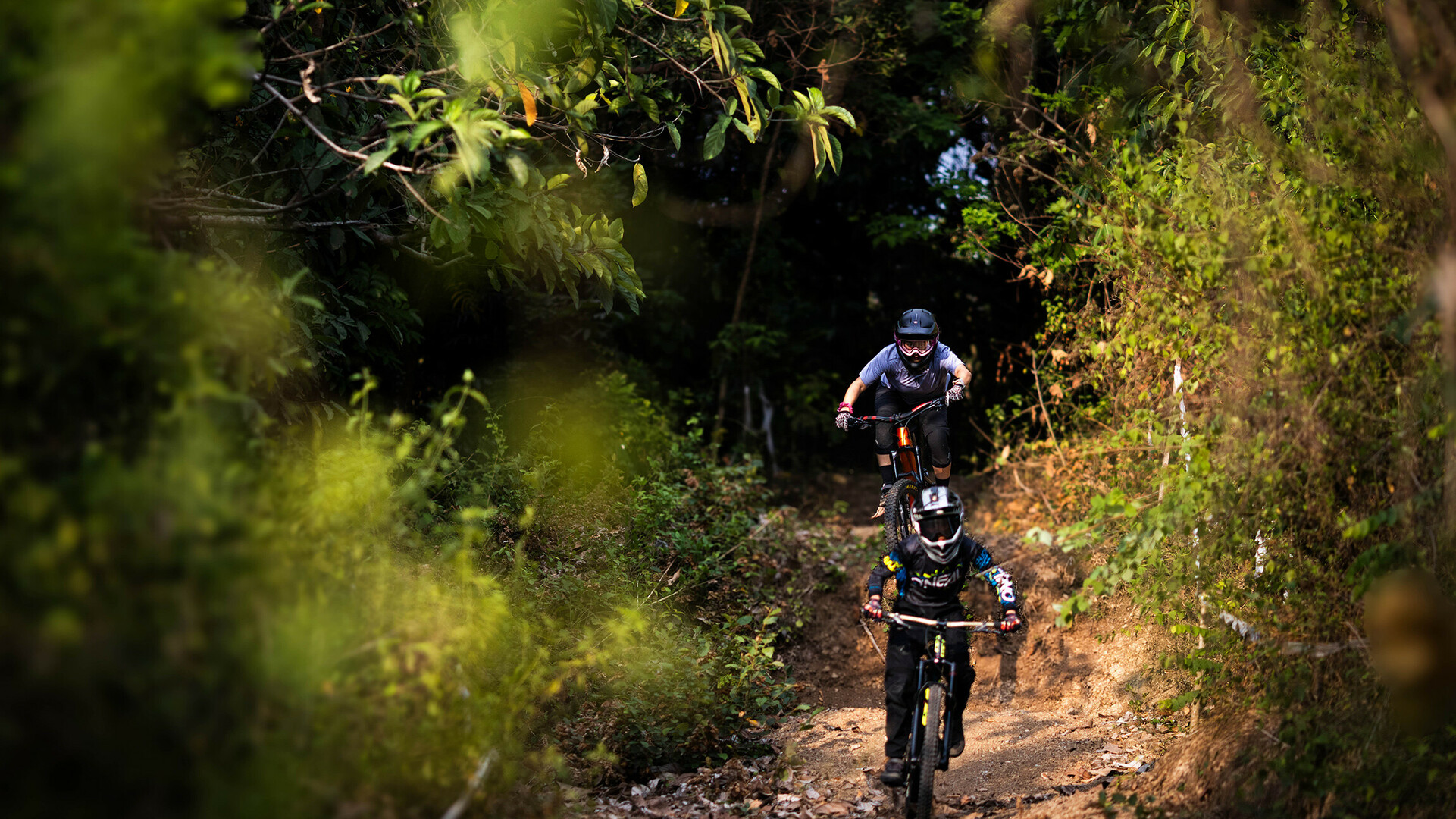 Zwei Mountainbiker mit Helmen fahren auf einer unbefestigten Straße durch einen Wald mit grünem Laub auf beiden Seiten.